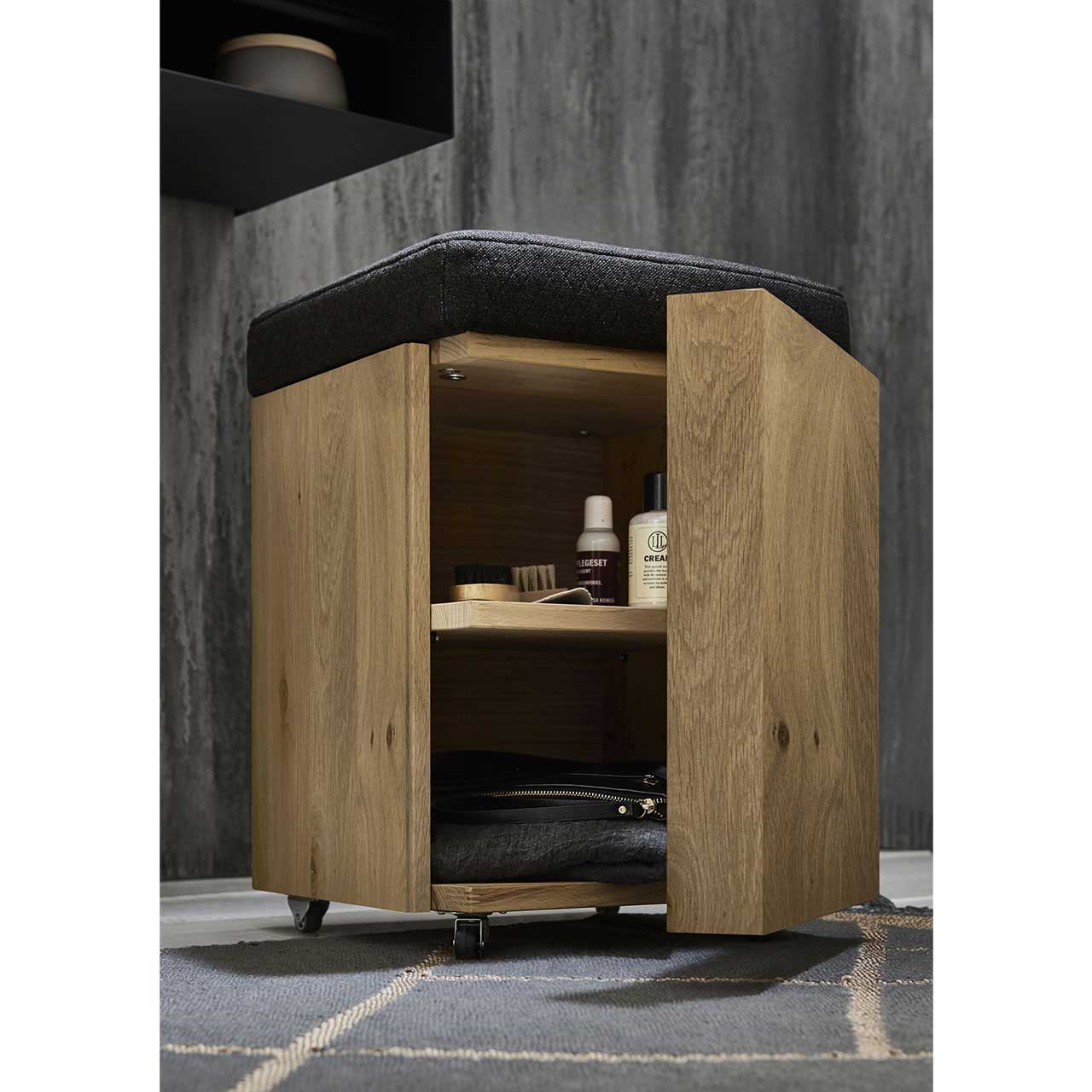 Praktischer Rollhocker aus massivem Holz mit Polstersitzfläche in Anthrazit und offener Holztür, dahinter sichtbar ein Holzeinlegeboden.