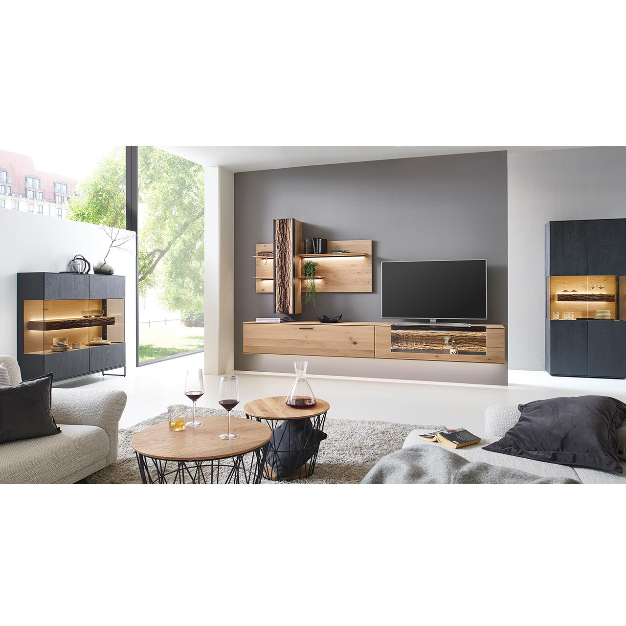 Wohnzimmer mit Wohnwand aus zwei Hängelowboards in massiver Kerneiche Natur, teils verglast, einem Wandpaneel mit beleuchteten Böden und Stauraumschrank, dazu links eine Standvitrine und rechts das passende Highboard in anthrazit gebeizter Kerneiche.