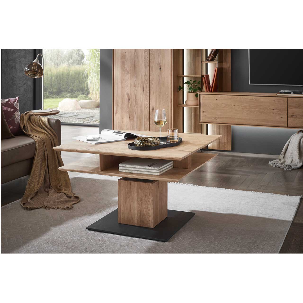 Moderner Couchtisch mit quadratischer, höhenverstellbarer Tischplatte aus hellem Eichenholz und zusätzlicher Holzablageplatte - auf massiver, quadratischer Holzsäule und dunkler Bodenplatte - im modern eingerichtetem Wohnzimmer stehend.