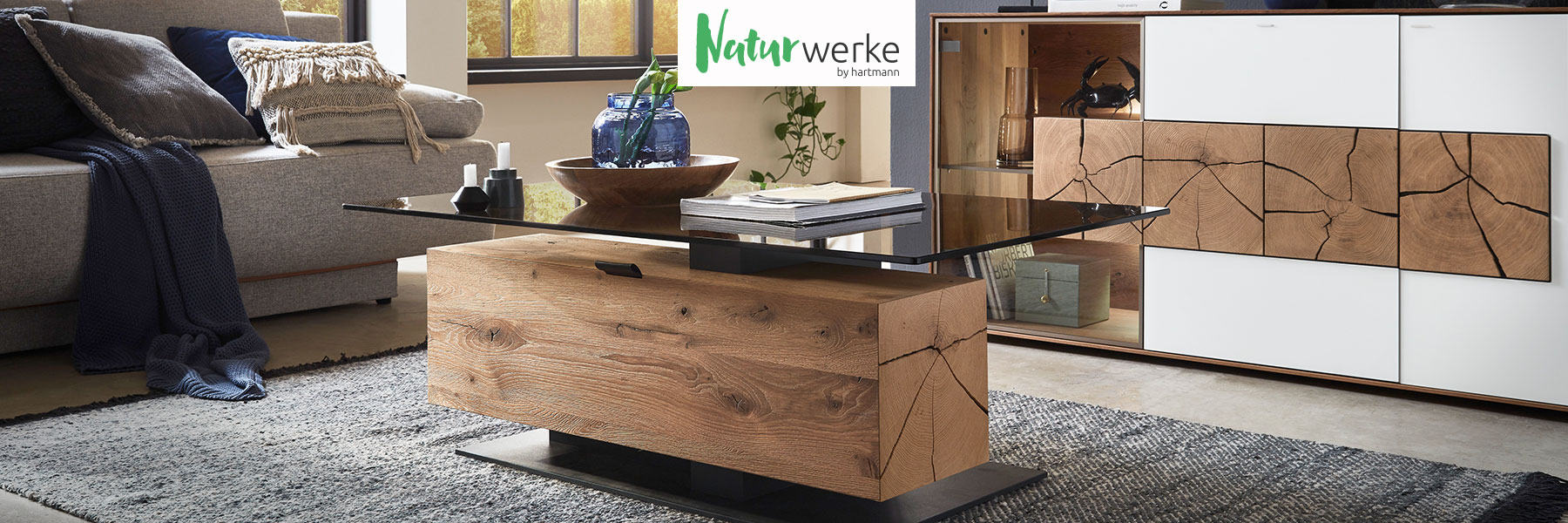 Hartmann Möbel - Möbelserie NATURWERKE - Kerneiche Natur oder Umato Hartmann NATURWERKE - individuelle und funktionelle Couchtische aus Massivholz kombiniert mit Glas und Metall. Ambientebild zeigt Couchtisch mit edler Glasplatte in Bronze und eckiger Holzsäule als Stauraumfach aus massiver Kerneiche Natur.