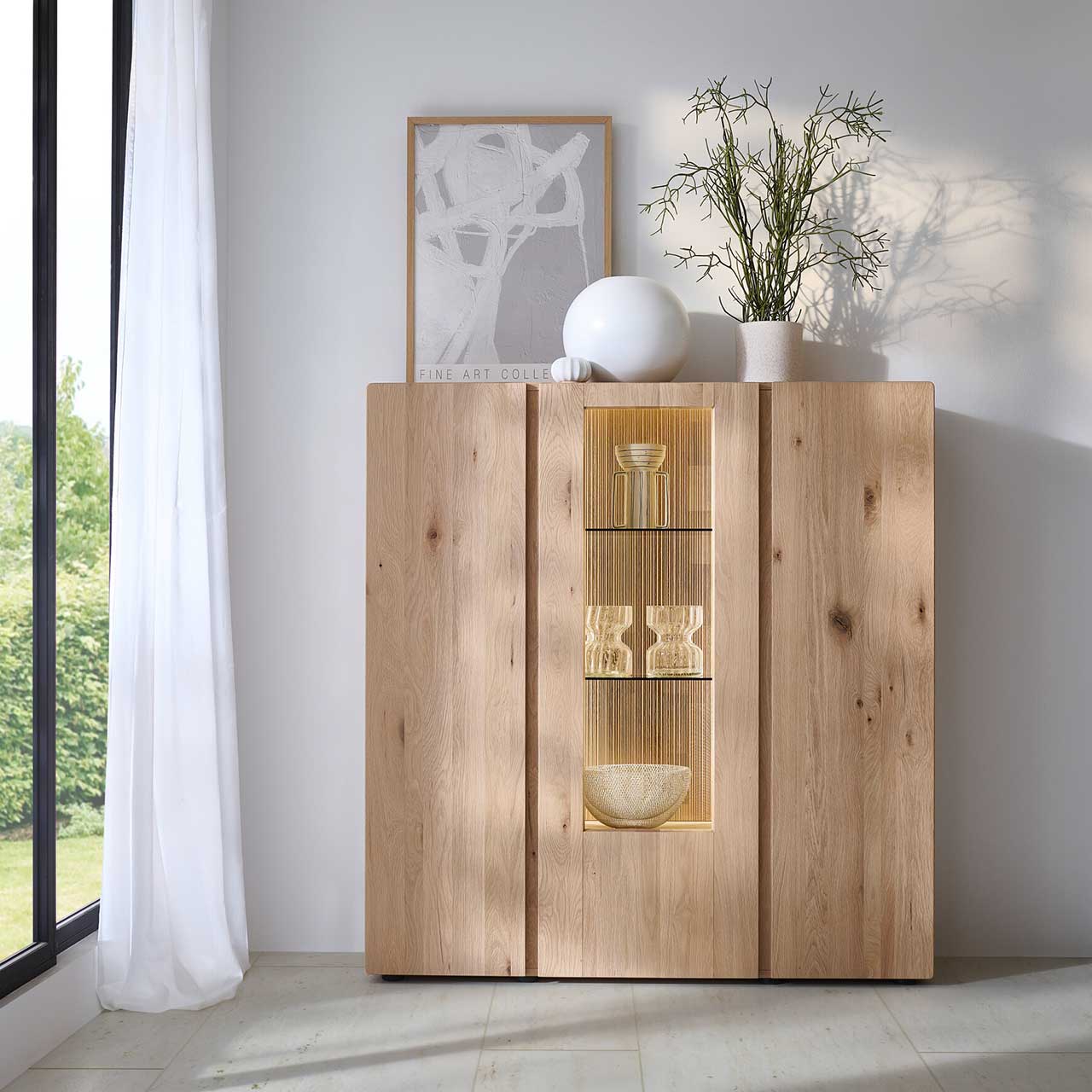 Modernes 3-türiges Highboard aus massiver Kerneiche Natur mit zwei Holztüren rechts und links und mittig mit einer beleuchteten Glas-/Holztür, dahinter sichtbar die edle geschoppte Rückwand - in Frontansicht an einer hellgrauen Wand stehend. 