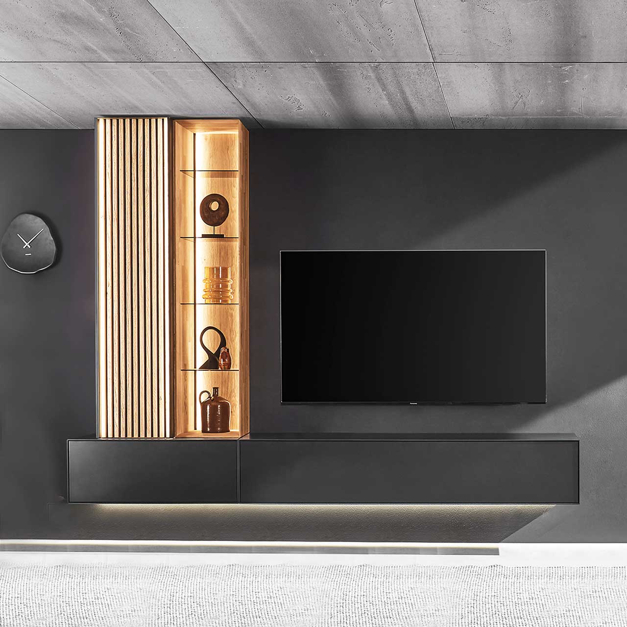 Hartmann SELINO - Wohnwand 8560 Nr. V26 Wohnzimmer mit Fernseher und moderner, vierteiliger Wohnwand aus massivem Holz: zwei anthrazit lackierte Hängelowboards, links darauf ein Hochschrank mit Lamellenfront und ein Wandregal mit Glasböden – vor dunkelgrauer Wand