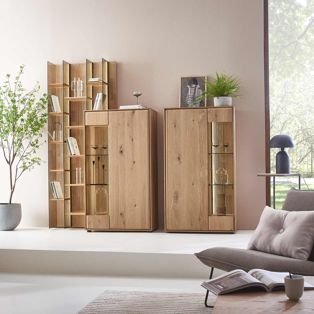 Zwei moderne Highboards aus massivem Eichenholz mit je zwei geschlossen Holz - und Glas/Holztüren. Links daneben in das Highboard integriert steht ein hohes Wandregal mit beleuchteten Holz und Glasböden - im Wohnzimmer stehend.