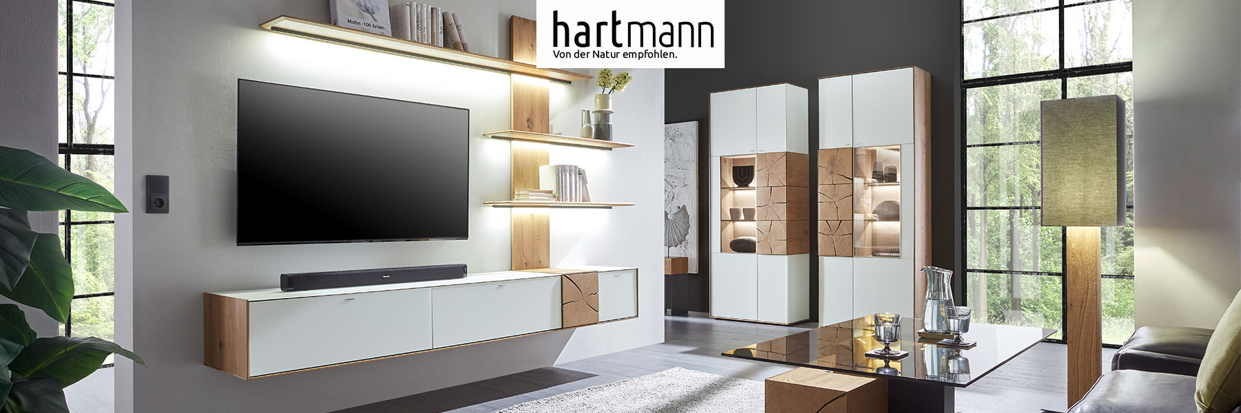 Hartmann Möbel - Möbelserie CAYA - Kerneiche Umato mit Mattglas weiß Hartmann CAYA - Massivholzmöbel aus Kerneiche Umato mit Mattglas weiß oder anthrazit Fronten und dekorativen Hirnholzscheiben. Ambientebild zeigt Hängelowboard weiß, beleuchtetes Wandpaneel aus Holz und zwei Standvitrinen mit Hirnholzscheiben.