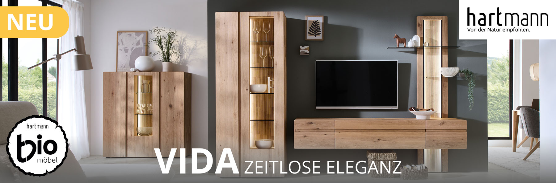 Hartmann VIDA - Massivholzmöbel aus Kerneiche Natur kombiniert mit geschroppten Rückwänden und Holzeinkerbungen.  Ambientebild zeigt Hängelowboard integriert in ein Wandpaneel mit Glasböden, links eine Standvitrine. Hinten links passendes Highboard.