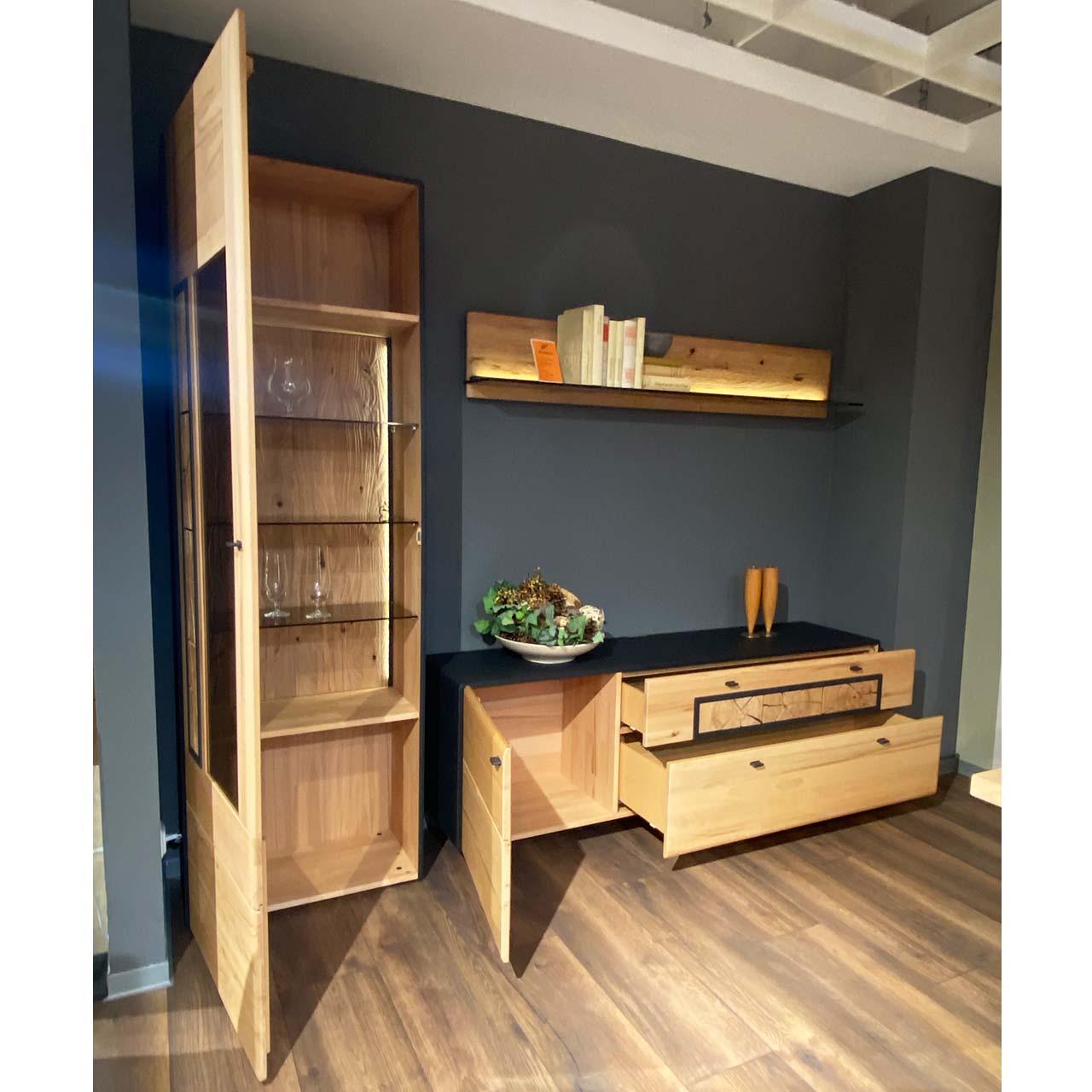 Dreiteilige Wohnwand aus massiver Kernbuche gebürstet: Hochvitrine mit geöffneter Holz/Glastür und Hirnholzscheiben mit drei Glaseinlegeböden, Lowboard mit offener Tür und zwei Schubkästen, darüber hängend ein beleuchtetes Wandpaneel.