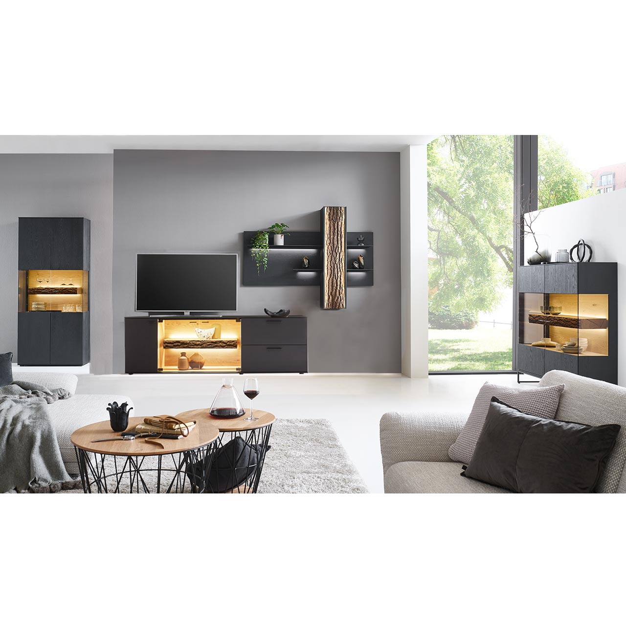 Wohnzimmer mit Wohnwand bestehend aus einem Lowboard in Kerneiche Natur anthrazit gebeizt mit Glasausschnitt und Rinden-Akzent und passendem Wandpaneel mit Böden und Stauraumschrank, dazu links Standvitrine und rechts Highboard.