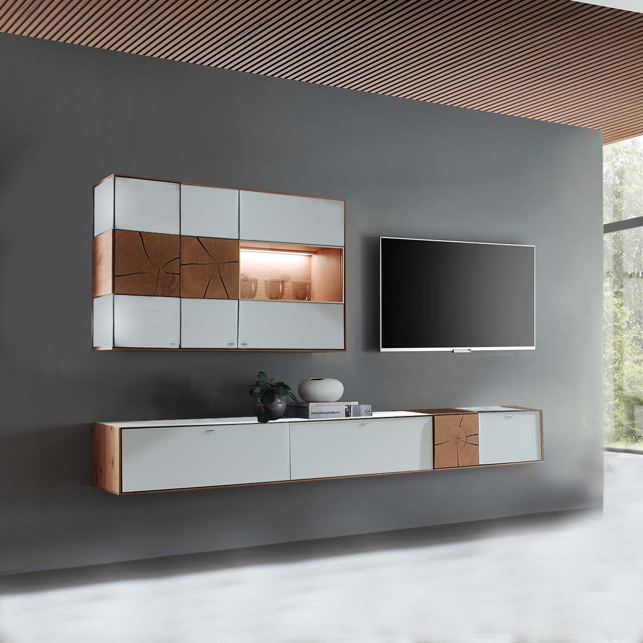 Hartmann CAYA - Wohnwand 7170 Nr. V76W - zweiteilig Massivholz Wohnwand aus Kerneiche mit Mattglas weißen Fronten. Unten ein langes, schmales Hängelowboard mit dekorativen Hirnholzscheiben, darüber die passende, beleuchtete Hängevitrine. Rechts daneben hängender Fernseher - vor dunkelgrauer Wand.