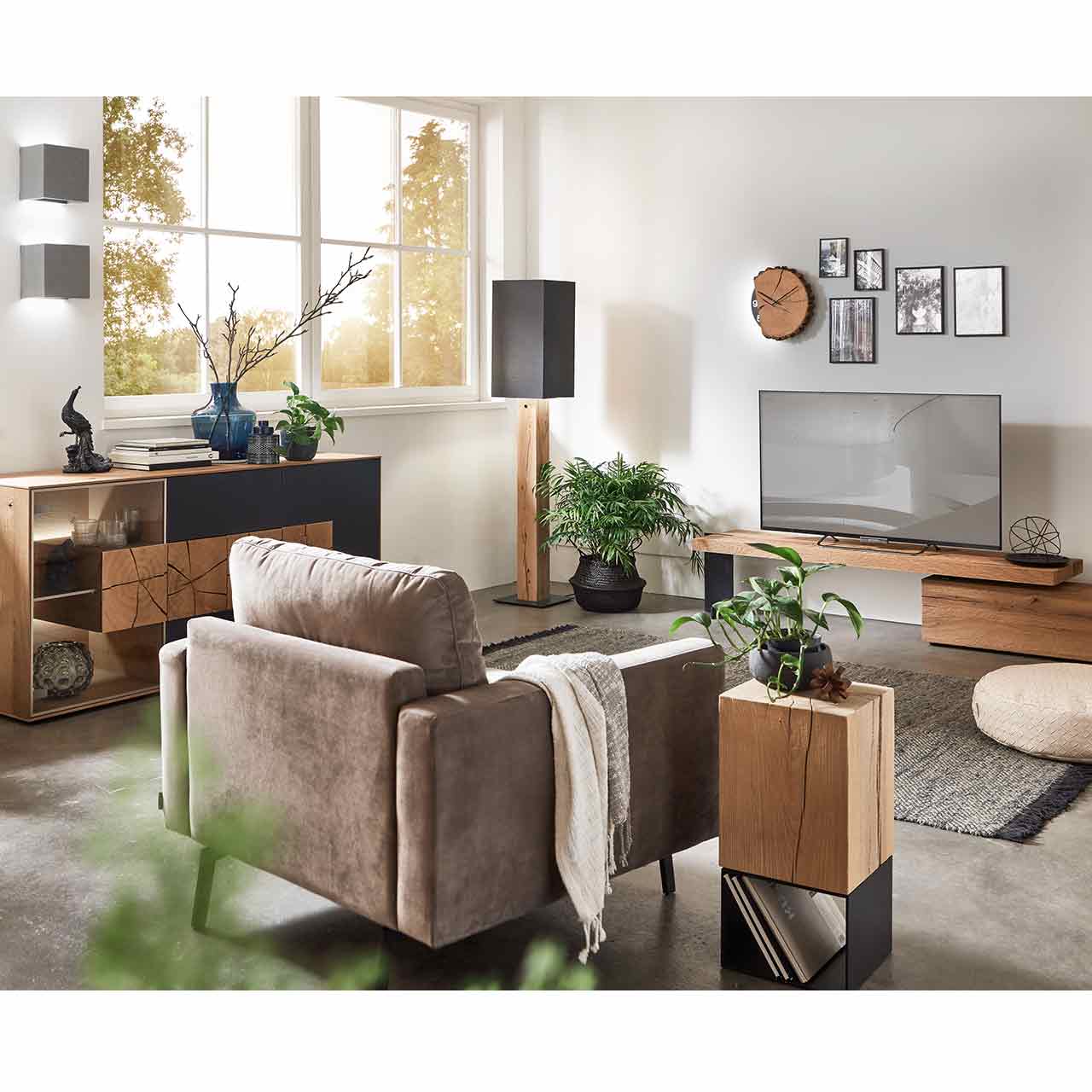 Eingerichtetes Wohnzimmer mit modernem Sideboard aus Kerneiche, Stehlampe mit Stofflampenschirm und Holzfuß und einer Holzwanduhr. Im Vordergrund ein passender Beistelltisch mit Holzkorpus und Metallgestell.