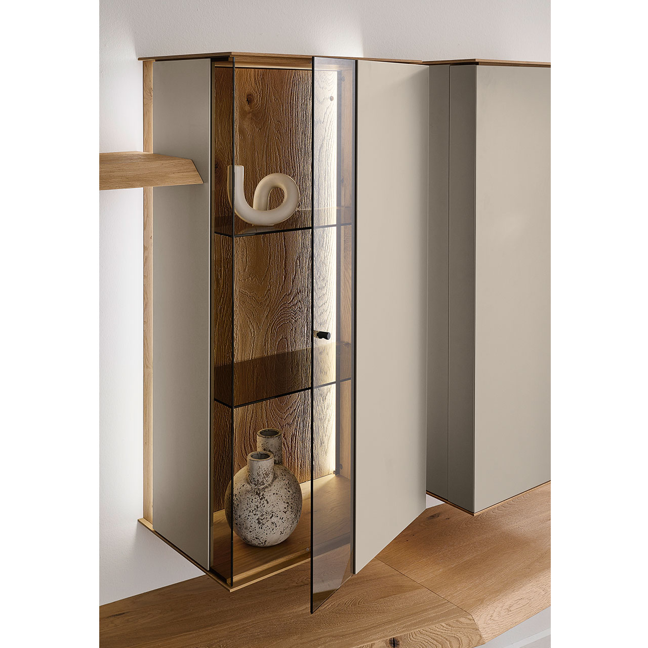 Detailansicht einer Vitrine aus massiver Kerneiche Natur mit Lackfronten in Sand. Mit geöffneter Glas-Lacktür, dahinter sichtbar die beleuchtete Riffholzrückwand sowie zwei Glasböden in Parsol Bronze.