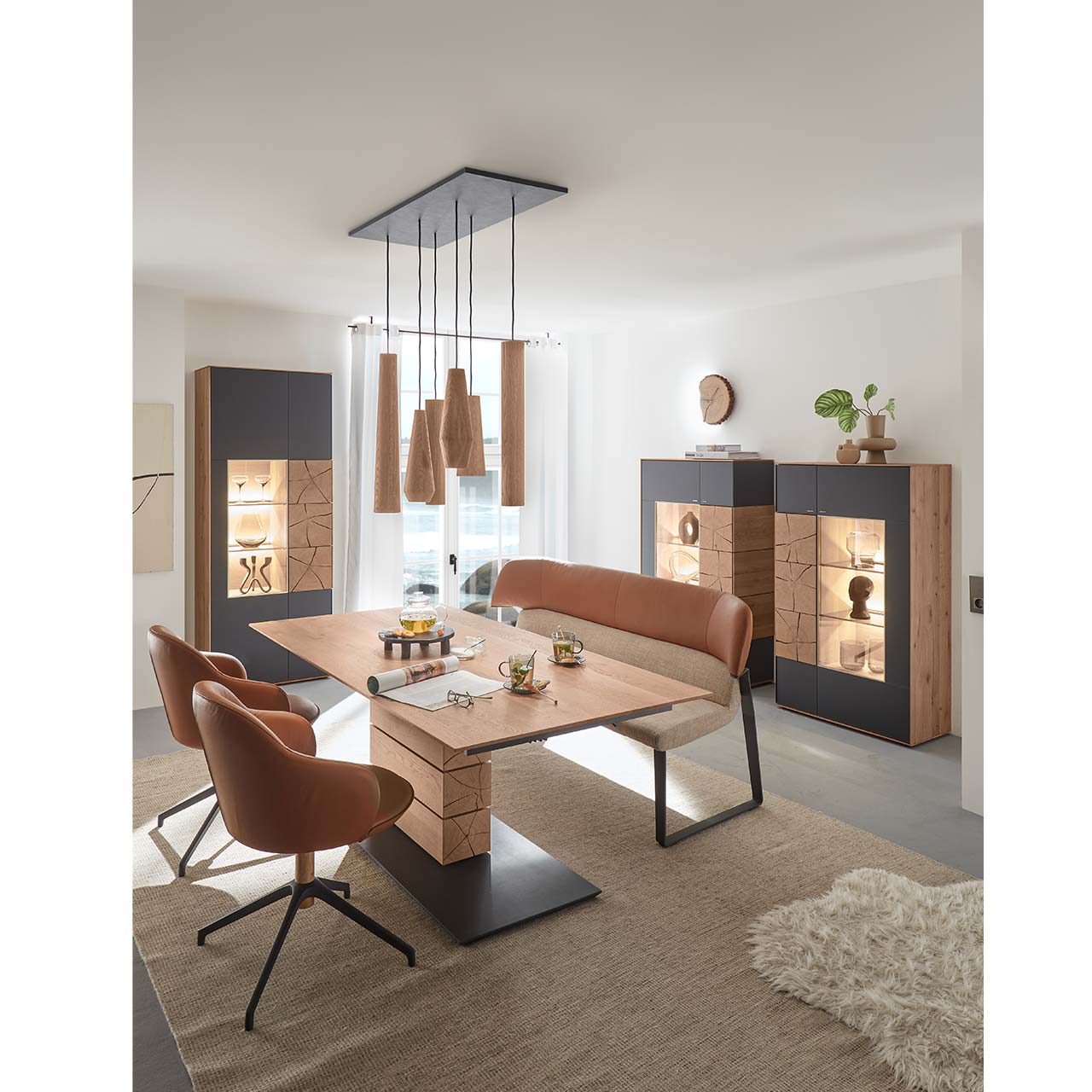 Modernes Esszimmer mit rechteckigem Massivholz Tisch mit Sitzbank und zwei Stühlen mit Lederbezug in cognac. Über dem Tisch hängt ein Pendelleuchte mit Holz-Lampenschirmen. Im Hintergrund stehen zwei passende Highboards und eine Standvitrine in Anthrazit.