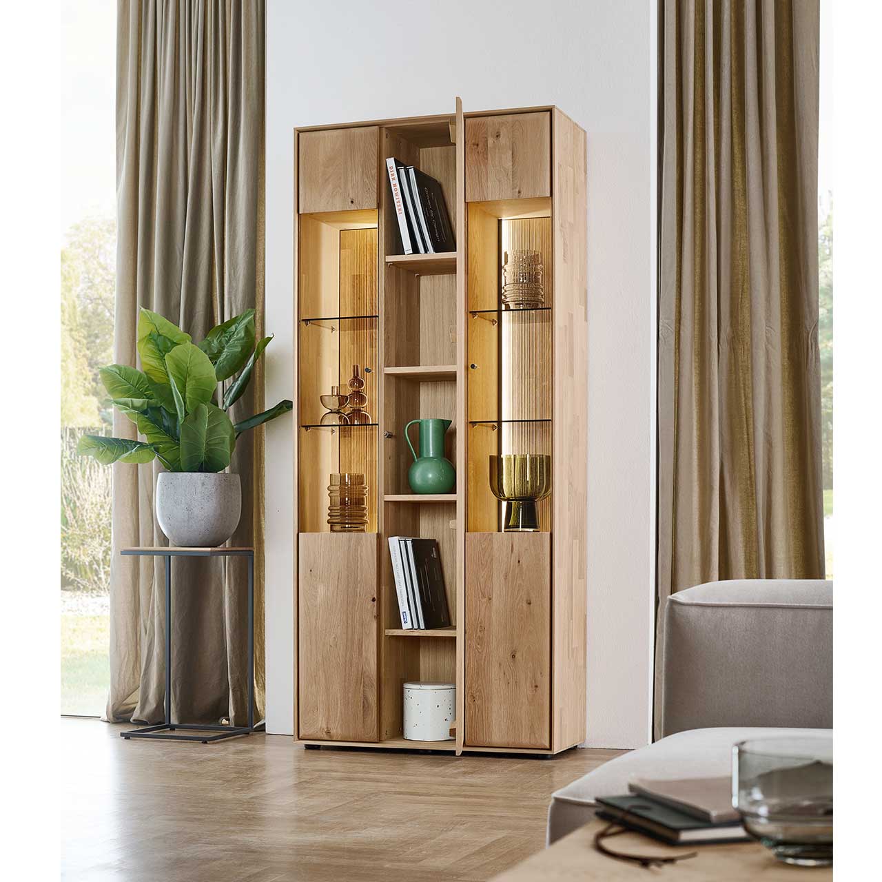 Moderne Standvitrine aus massivem Eichenholz mit 2 geschlossenen, beleuchteten Glastüren rechts und links und einer geöffneten Holztür mittig, darin sichtbar 4 Holzeinlegeböden mit Deko - im Wohnzimmer stehend.