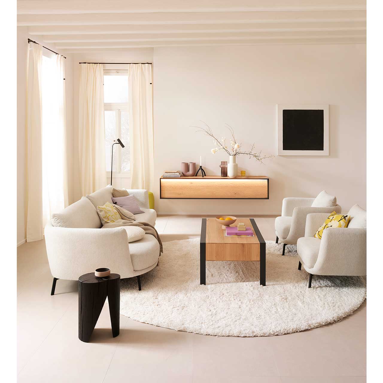Helles Wohnzimmer mit beleuchtetem Hängelowboard aus massiver sandgestrahlter Kerneiche Natur eingefasst in Metallrahmen anthrazit an hellbeiger Wand. Davor steht ein rechteckiger Holz-Couchtisch auf Metallfüßen anthrazit.
