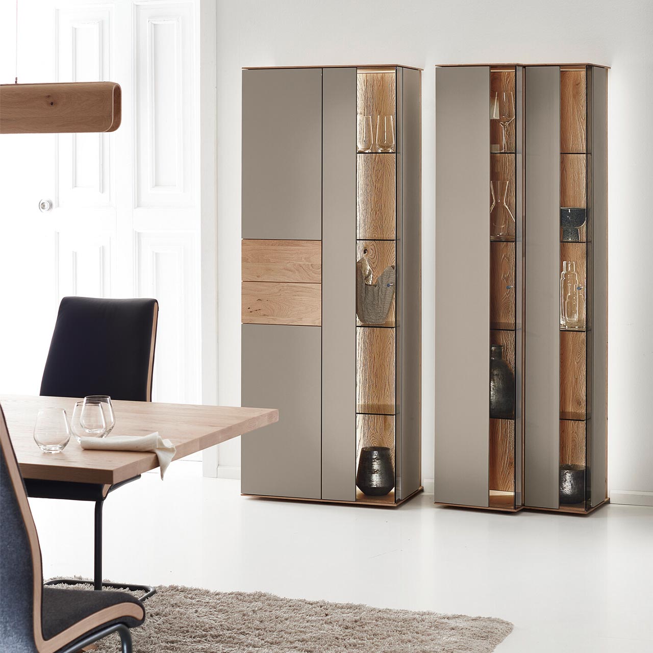 Moderne Standvitrinen aus massiver Kerneiche Natur mit Lackfronten in Sand. Mit edlen Glastüren in asymmetrischem Design und  beleuchteten Riffholzrückwänden.