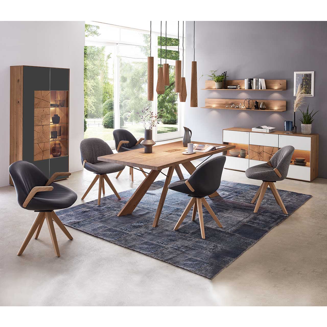 Esszimmer mit massivem Holztisch und fünf Stühlen mit Holzgestell, links an der Wand stehend eine Strandvitrine in Mattglas anthrazit und beleuchtetem Glasausschnitt mit drei Glasböden. Rechts das passende Sideboard und zwei Wandregale.