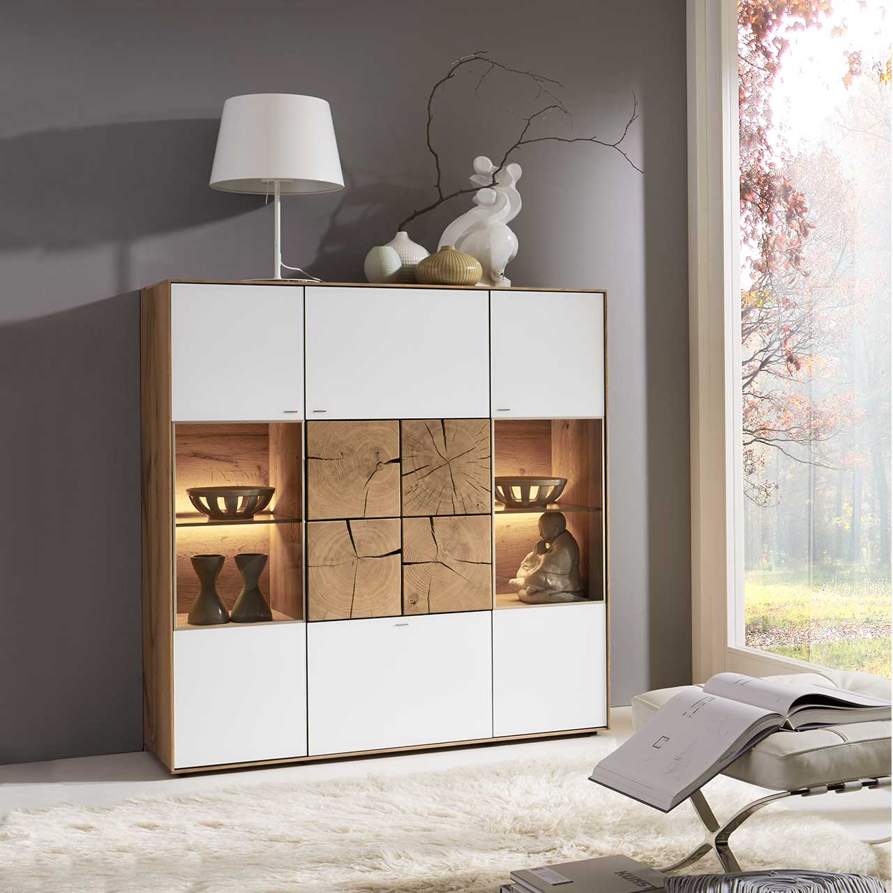 Milieubild Hartmann CAYA - Highboard 7170-7138W Modernes Highboard aus massiver Kerneiche Umato mit Mattglas weiß Fronten, rechts und links mit beleuchtetem Glasausschnitt, mittig mit vier dekorativen, rechteckigen Hirnholzscheiben in heller Kerneiche Umato - in einem modernen Flur stehend.