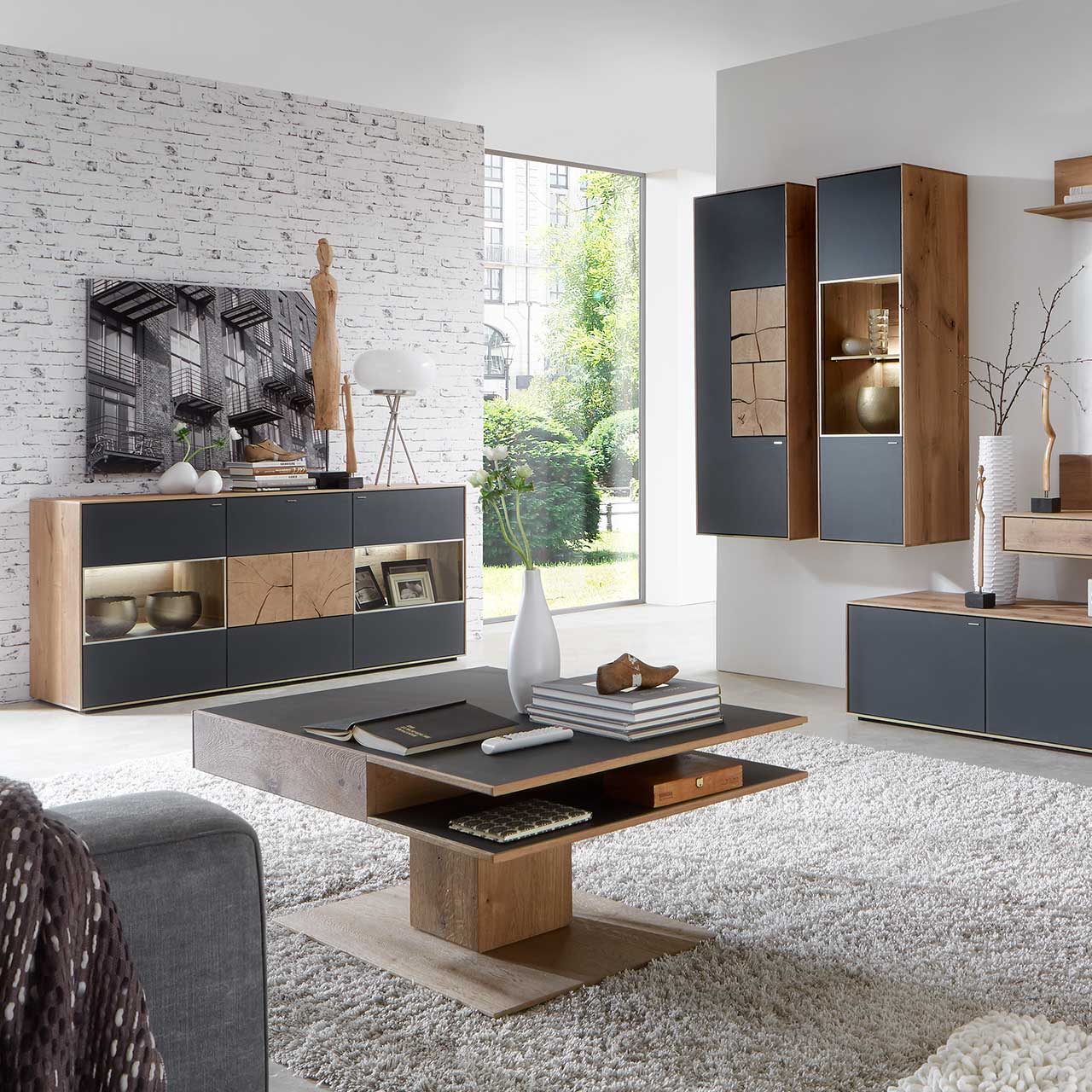 Wohnzimmer mit modernen Massivholzmöbel aus Kerneiche Umato mit Mattglas anthrazit Fronten: Hängeschrank mit Hirnholzscheiben, beleuchtete Hängevitrine, links Sideboard mit Glasfront und Hirnholzscheiben, davor rechteckiger Holz-Couchtisch mit Ablagefach.