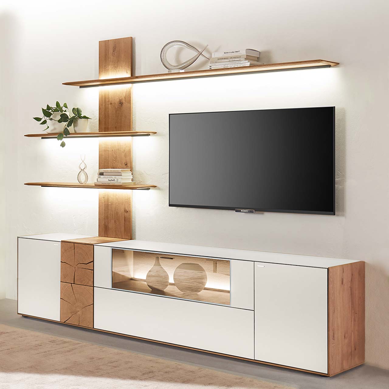 beleuchteten Regalböden. Dazwischen hängender Fernseher. Hartmann CAYA - Wohnwand 7170 Nr. V80W - zweiteilig Moderne zweiteilige Massivholz Wohnkombination aus Kerneiche Umato mit Mattglas weiß Fronten und Hirnholzscheiben, mit einem langen Lowboard mit Glasausschnitt und einem Wandpaneel mit drei beleuchteten Regalböden. Dazwischen hängender Fernseher.