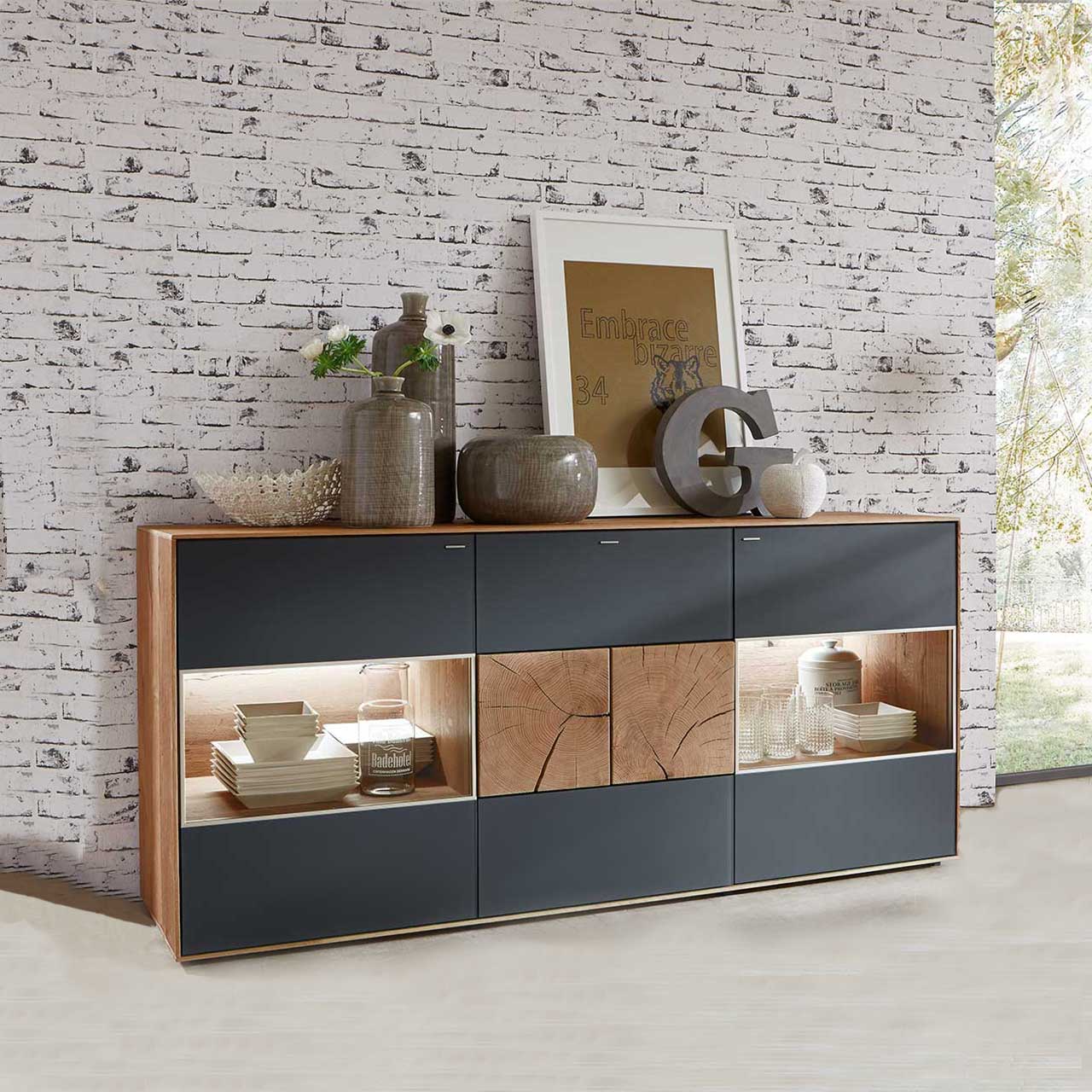Modernes Sideboard aus massiver Kerneiche Umato mit anthrazitfarbenen Mattglasfronten. Rechts und links mit beleuchtetem Glasausschnitt, mittig mit zwei dekorativen, rechteckigen Hirnholzscheiben - vor hellgrauer Steinwand-Tapete.