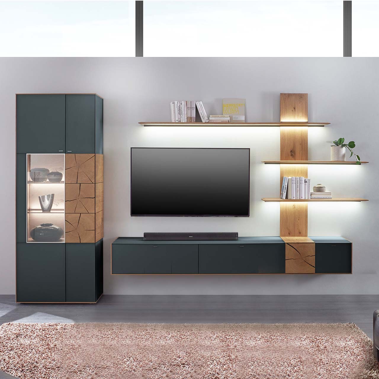 Hartmann CAYA - Wohnwand 7170 Nr. V74A - dreiteilig Massivholz Wohnwand aus Kerneiche mit anthrazitfarbenen Fronten. Unten ein Hängelowboard mit dekorativer Holzapplikation, seitlich ein Wandpaneel mit Regalböden und indirekter Beleuchtung, links passende Standvitrine, dazwischen hängender Fernseher.