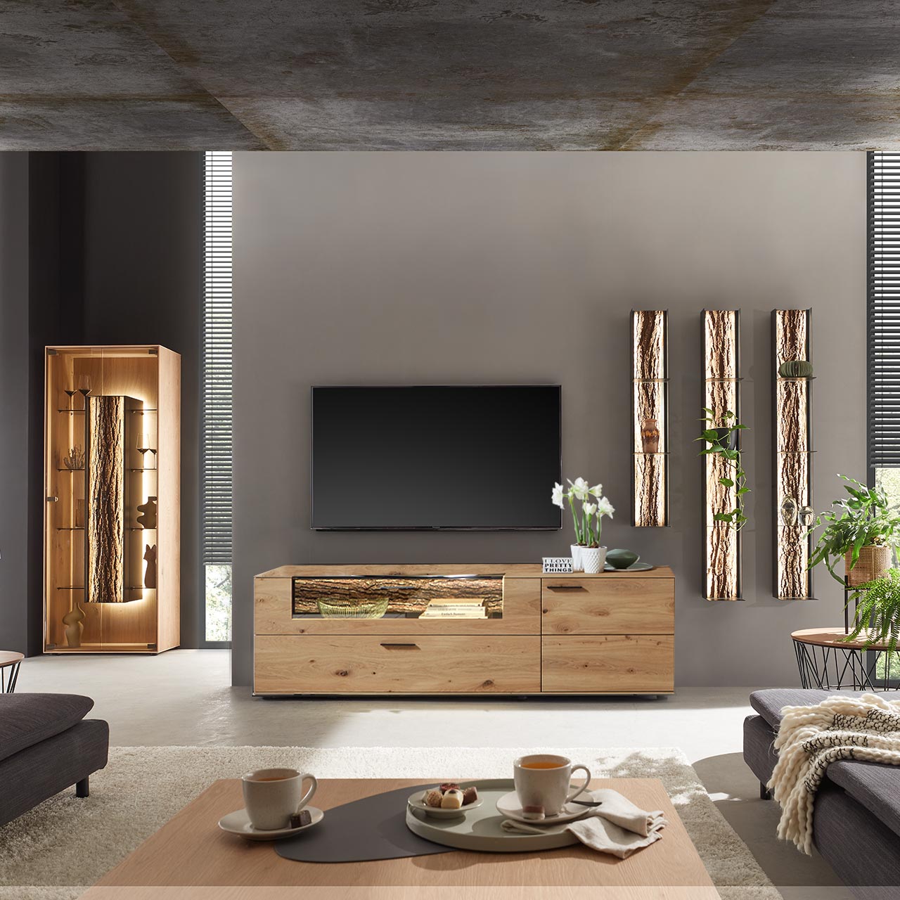 Modernes Wohnzimmer mit Wohnwand bestehend aus einem Lowboard aus massiver Kerneiche Natur, mit beleuchtetem Glasausschnitt und Rinden-Akzent und drei modernen Wandregalen aus Metall mit sichtbarer Baumrinde. Links die passende Standvitrine.