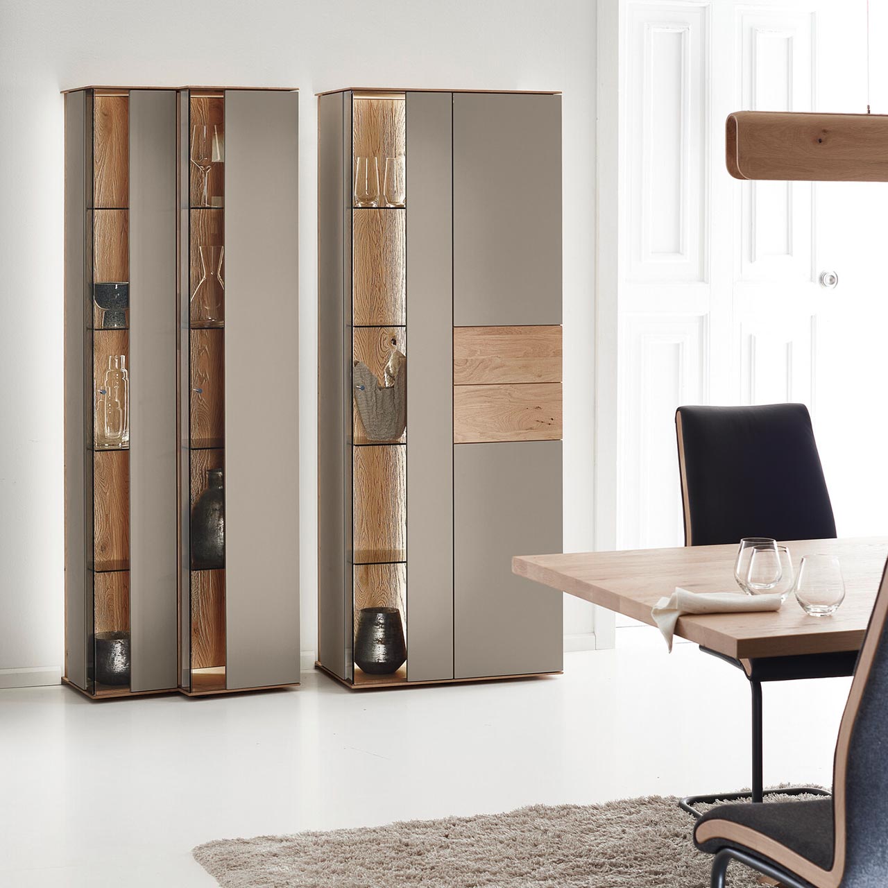 Moderne Standvitrinen aus massiver Kerneiche Natur mit Lackfronten in Sand. Mit edlen Glastüren in asymmetrischem Design und  beleuchteten Riffholzrückwänden.