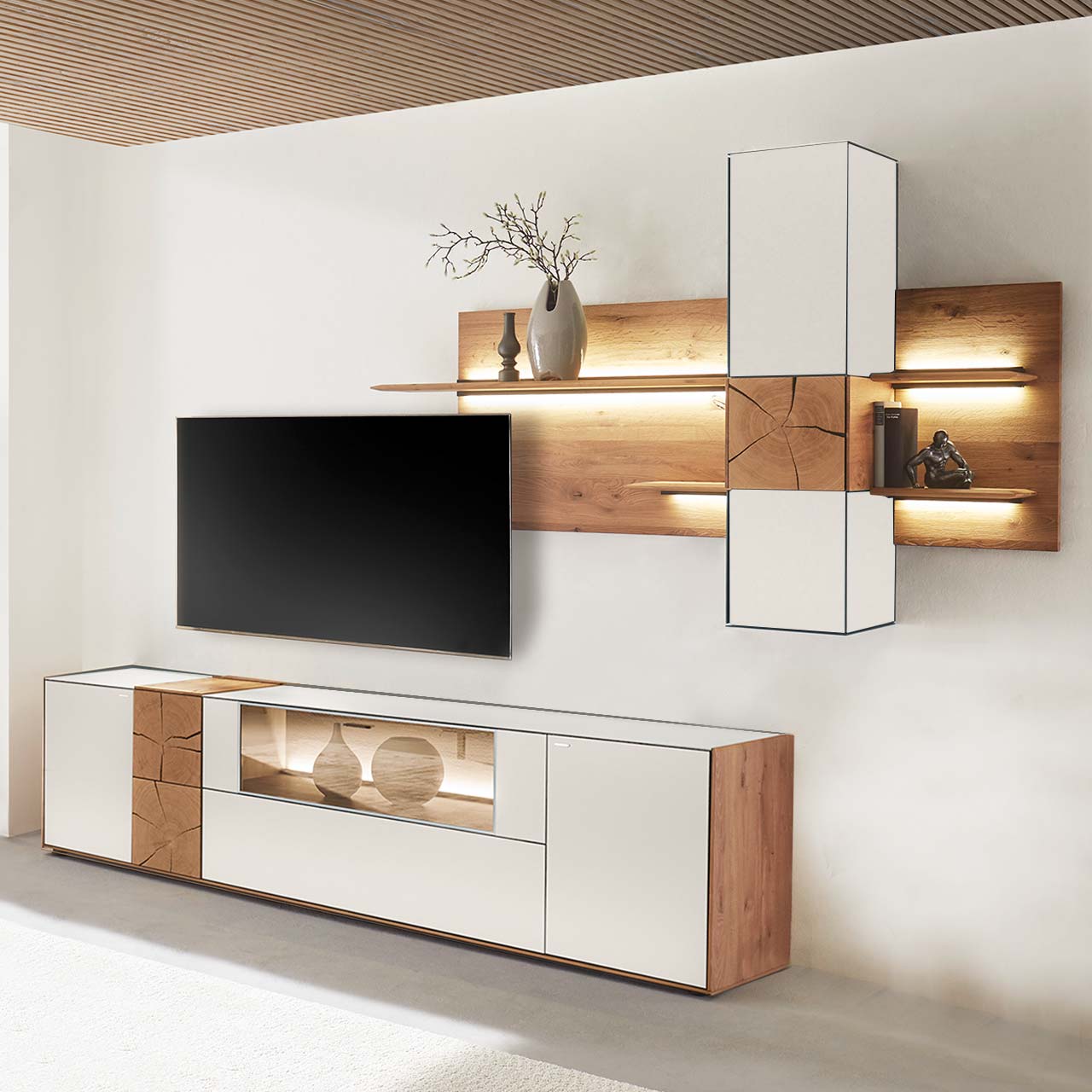 Hartmann CAYA - Wohnwand 7170 Nr. V70W - dreiteilig Moderne Wohnwand aus massiver Kerneiche mit Mattglas weißen Fronten. Unten ein Lowboard mit Vitrine und dekorativer Holzapplikation, oben eine Wandpaneel-Kombination mit Regalböden, Schrank und indirekter Beleuchtung, dazwischen hängender Fernseher.