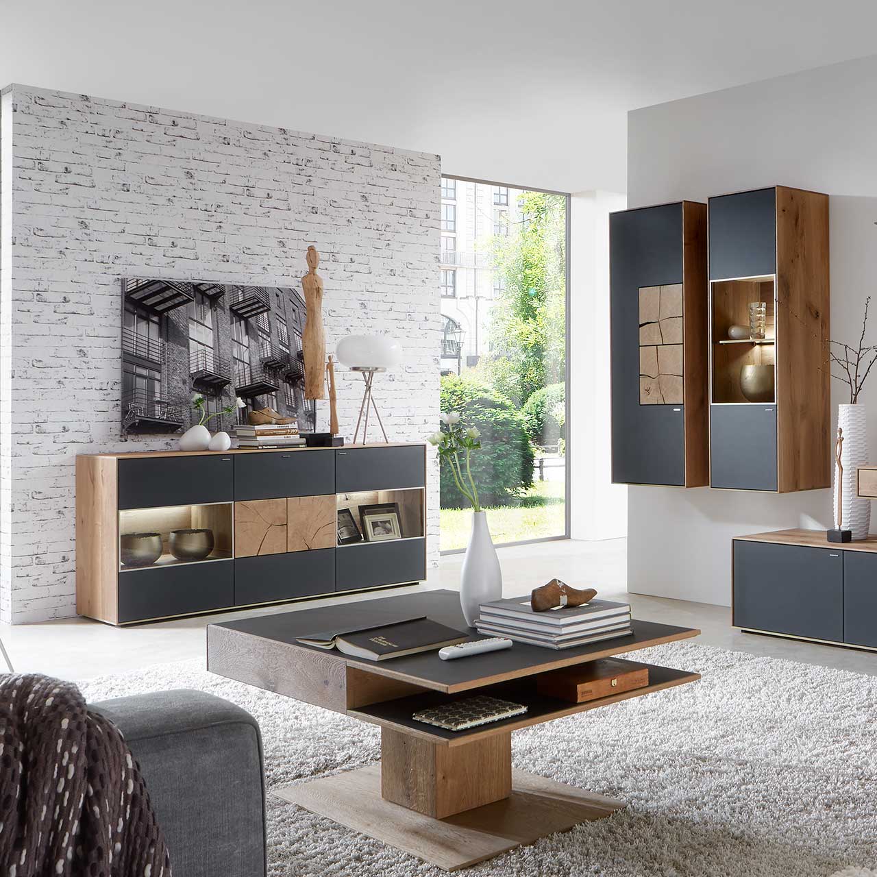 Wohnzimmer mit modernen Massivholzmöbel aus Kerneiche Umato mit Mattglas anthrazit Fronten: Hängeschrank mit Hirnholzscheiben, beleuchtete Hängevitrine, links modernes Sideboard mit Glasfront und Hirnholzscheiben, davor passender Couchtisch.