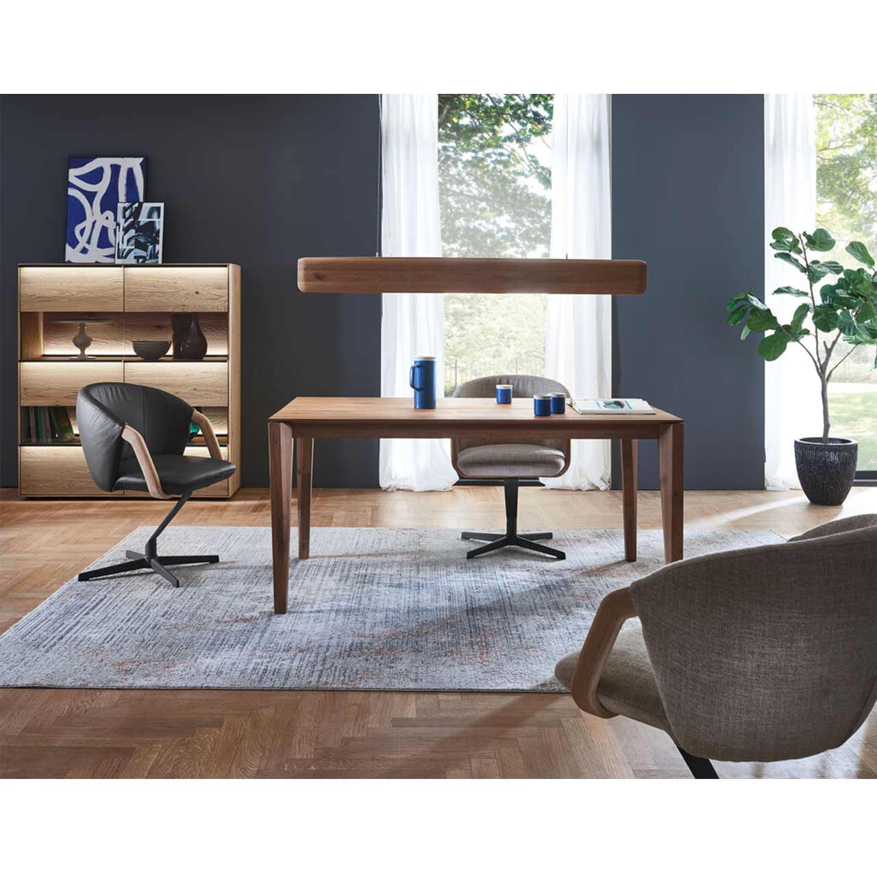 Esszimmer mit klassischem Holztisch aus massiver Kerneiche und drei modernen Stühlen mit Metallgestell. Darüber hängend eine Balkenleuchte. Links vor dunkelgrauer Wand das passend Highboard mit beleuchteter Glasfront.  