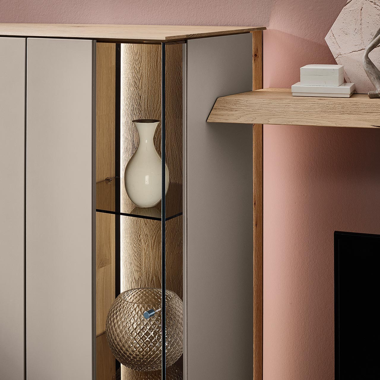 Detailansicht einer Vitrine aus massiver Kerneiche Natur mit Lackfronten in Sand mit Glas-Lacktür, dahinter sichtbar die beleuchtete Riffholzrückwand sowie zwei Glasböden in Parsol Bronze.