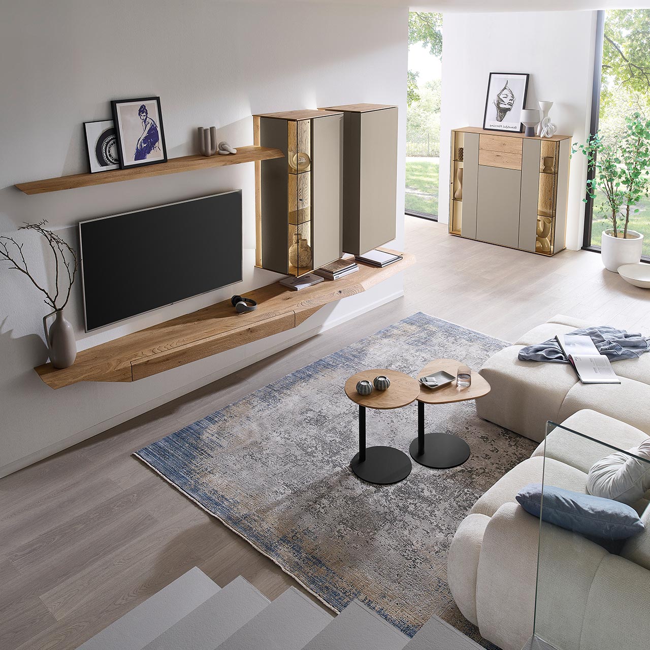 Wohnzimmer mit moderner Massivholzwohnwand bestehend aus Hängelowboard, Hängevitrine, Hängeschrank sowie Wandboard - im asymmetrischen Design. Rechts passendes Highboard mit Glastüren, davor zwei Beistelltische aus Holz - Ansicht von oben.