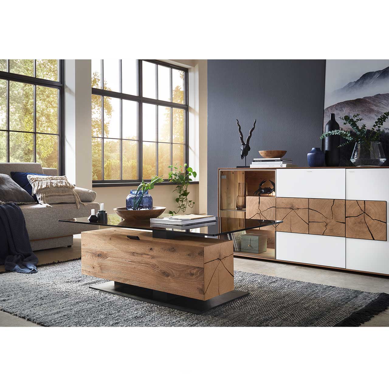 Moderner Couchtisch mit rechteckiger Glasplatte und Hirnholzblock aus massiver Kerneiche mit Klappe und Stauraumfach auf dunkler Bodenplatte. Dahinter ein Sideboard mit weißer Front und Hirnholzscheiben - im Wohnzimmer stehend. 