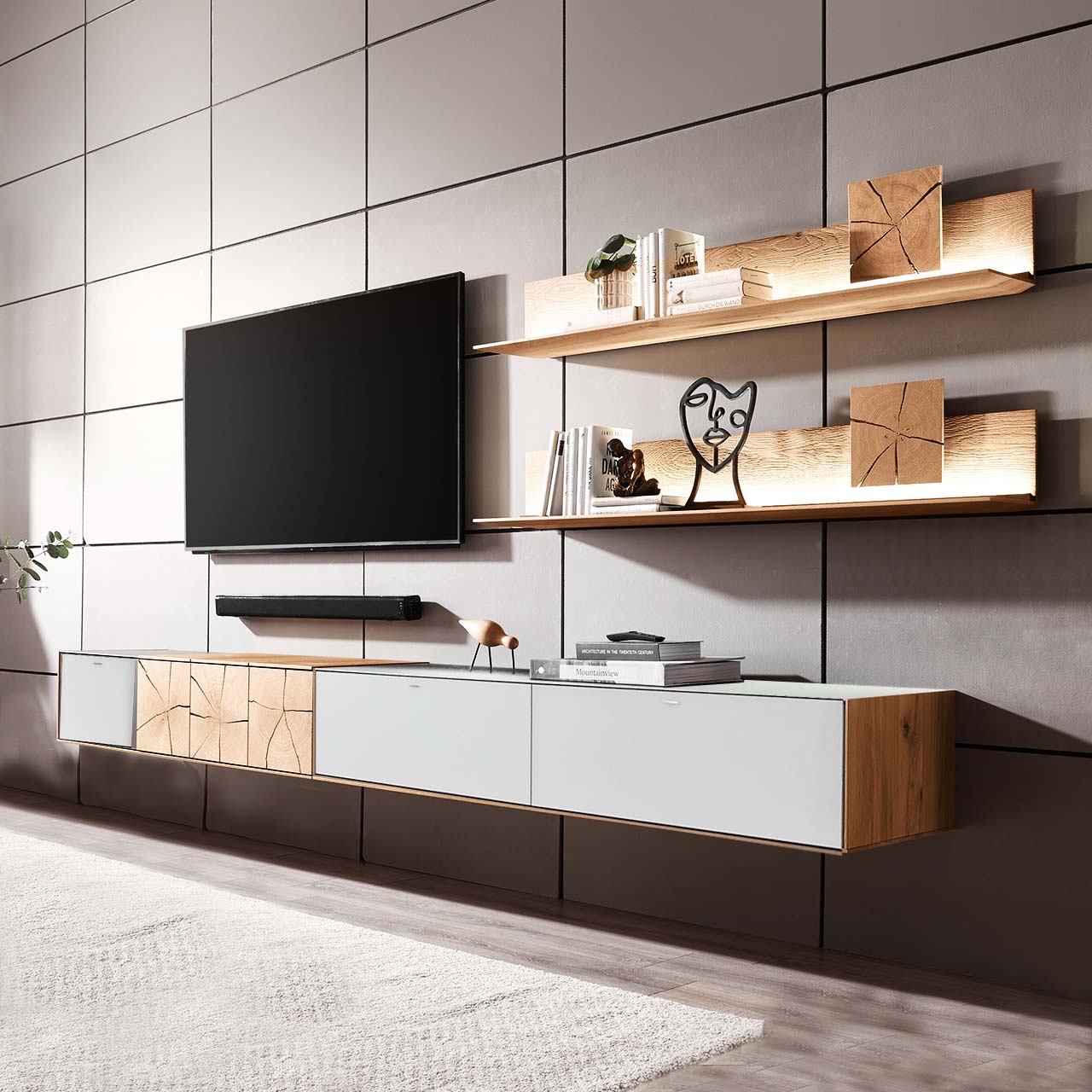 Hartmann CAYA - Wohnwand 7170 Nr. V90W - zweiteilig Moderne zweiteilige Massivholz Wohnkombination aus Kerneiche Umato mit Mattglas weiß Fronten, mit langem Hängelowboard und zwei Wandpaneelen mit dekorativen Hirnholzscheiben. Links daneben hängender Fernseher.